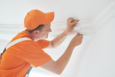 An image of a contractor, dressed in orange, installing white moulding onto a white wall.