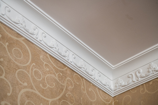 An image of decorative, white crown moulding on a wallpapered wall and a tan ceiling.